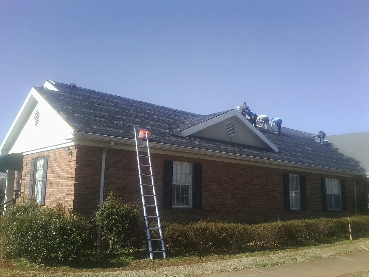 Crew tearing shingles off a brick ranch roof with a ladder set — Metal Roof Repair work by our crew in Drummond