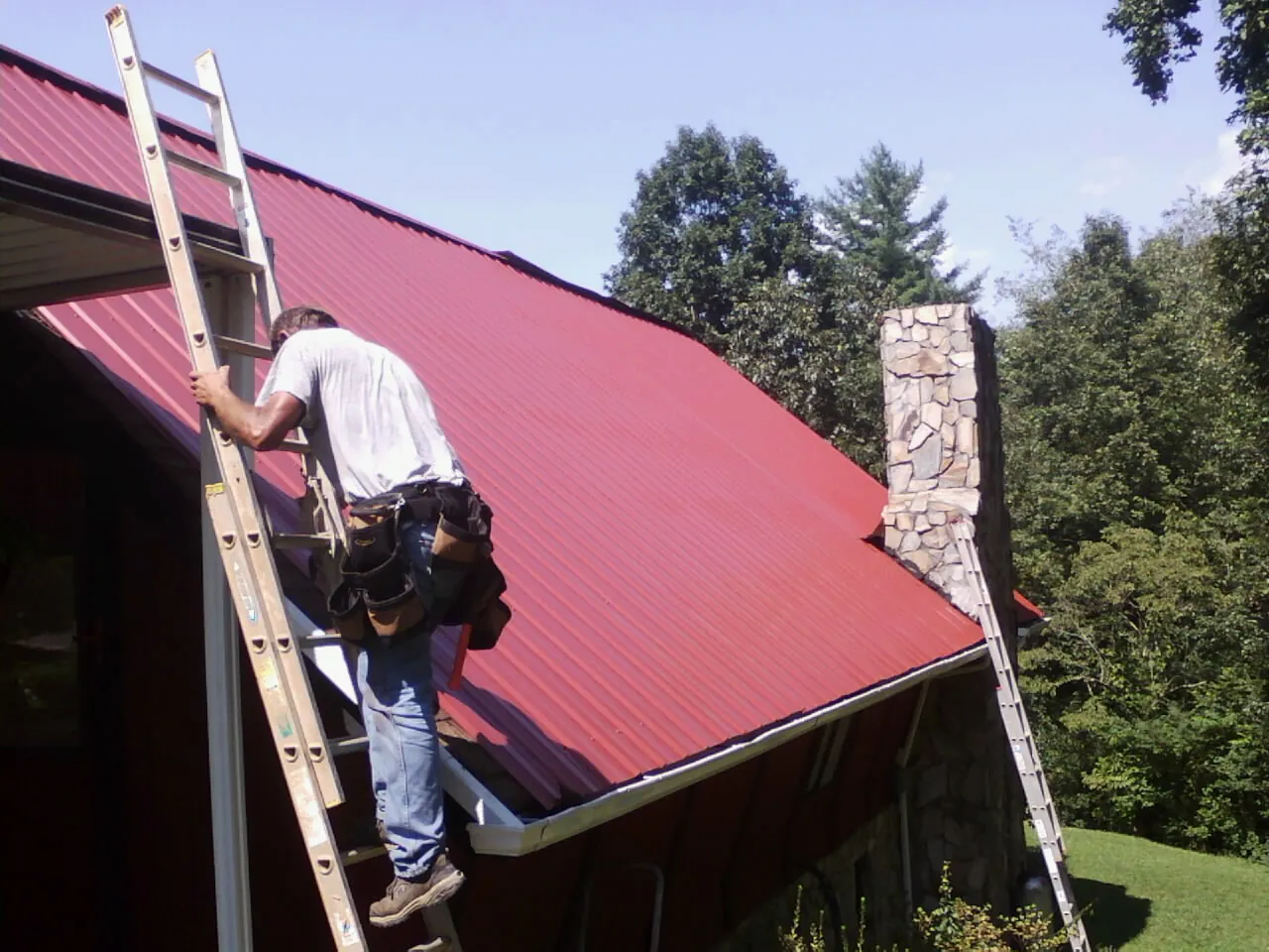 Roofer on a ladder beside a red metal roof and stone chimney — Soffit & Fascia Repair by Roofing Brothers in Drummond
